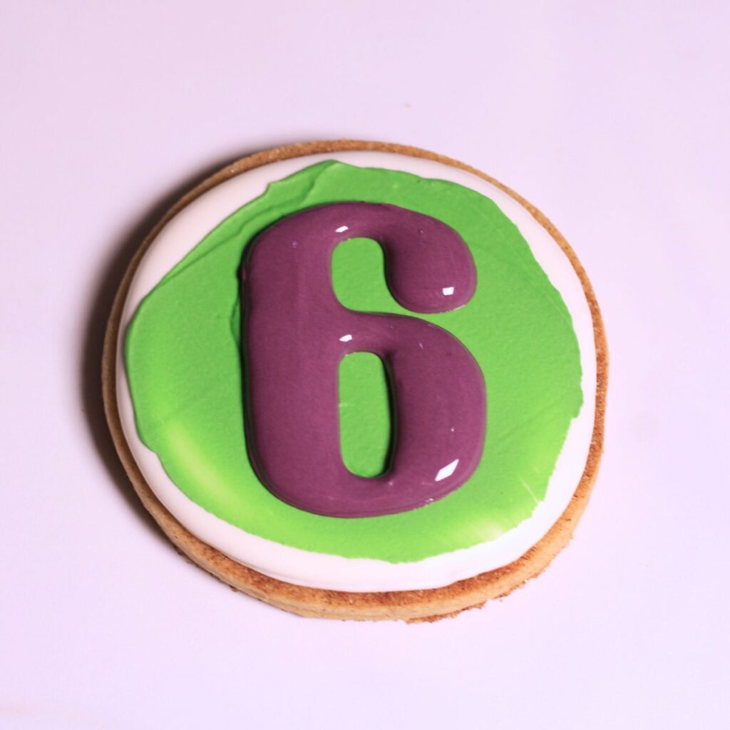 A circular cookie with green and white icing and a purple number 6 on the top center.