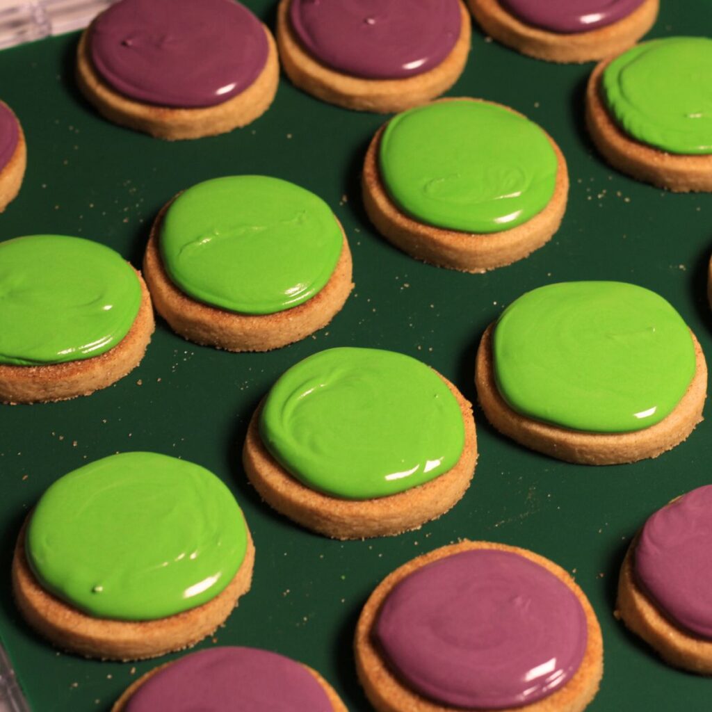 A tray of mini circle cookies: some covered in bright green icing the others in purple icing.