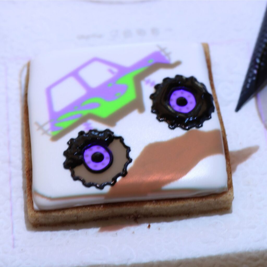 A square cookie covered in white icing with a monster truck projected onto the center. Black icing covers the projected tires.