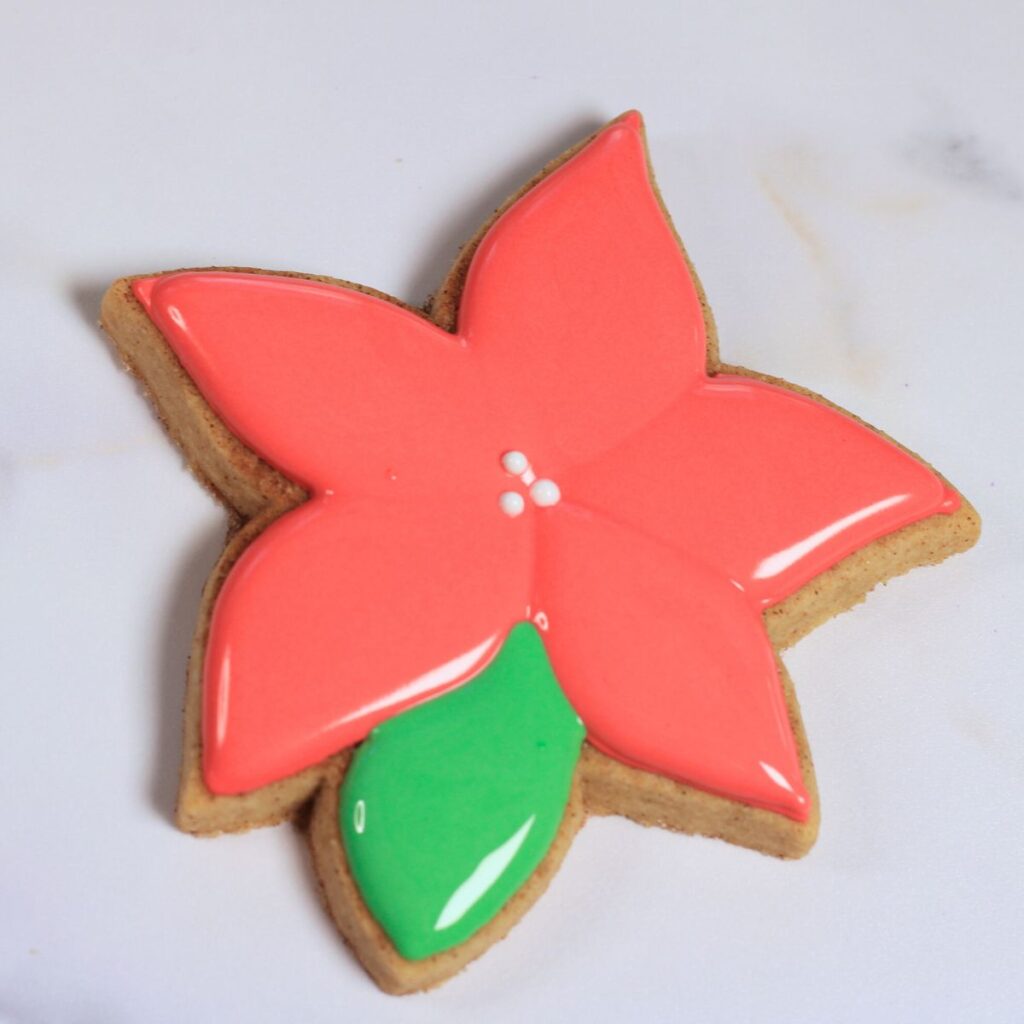 Poinsettia flower shaped cookie with five red petals, one green leaf, and three white dots in the center.