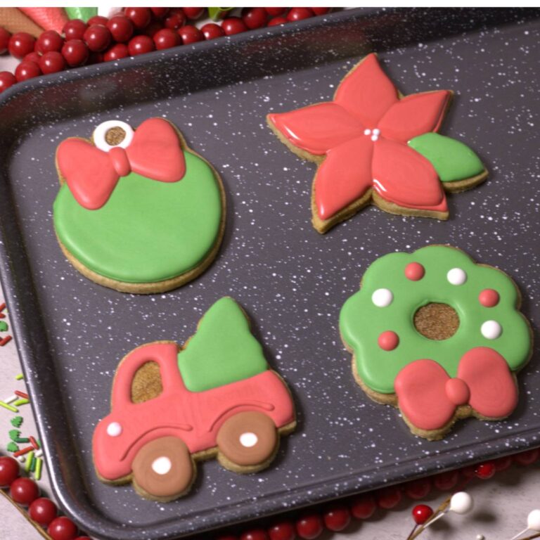 Christmas Cookies on a grey speckled tray: a green ornament with red bow, a red poinsettia flower with green leaf, a red truck with a green Christmas tree in the bed, and a green wreath with a red bow and red and white ornaments on the foilage.
